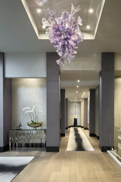A modern hotel lobby or hallway with a glass chandelier hanging from a recessed ceiling, featuring purple and clear glass elements. The space has gray columns, neutral-colored walls, a decorative console table with a white floral arrangement and a sm