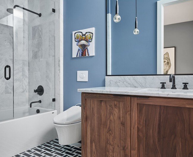 Modern bathroom with blue wall, marble countertop, wooden vanity, framed artwork, and two hanging light bulbs.