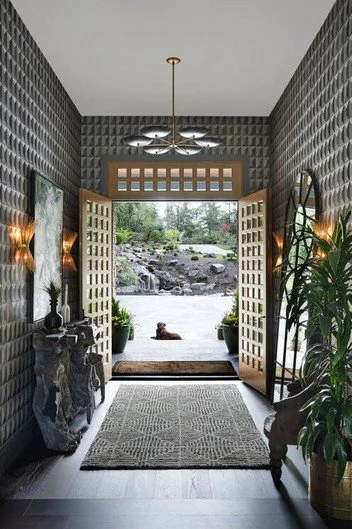 Interior view of a modern entryway with open wooden doors leading outside to a river and rocks, decorated with plants, wall-mounted lamps, a patterned rug, and contemporary lighting fixture.