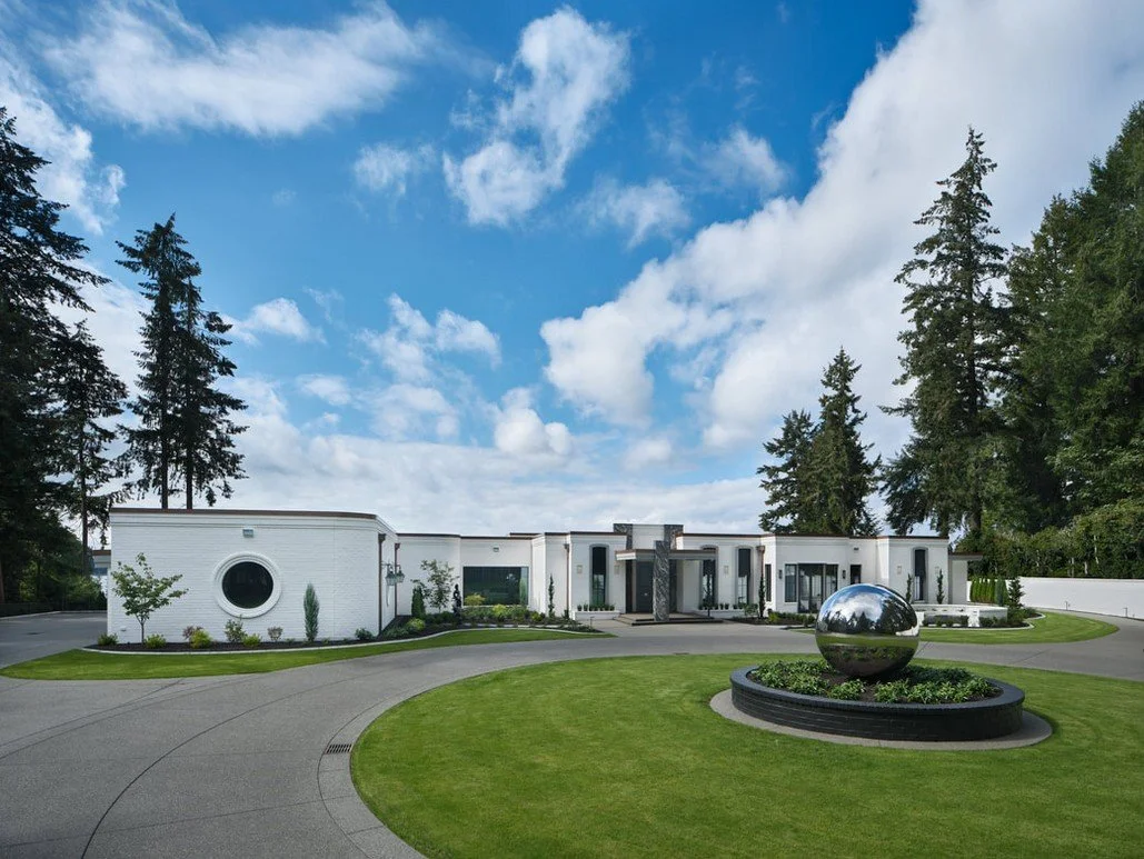 Modern white house with curved driveway, surrounded by green lawn and trees, and a shiny reflective sculpture in the front yard under a blue sky with clouds.