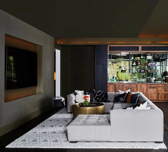 Modern living room with a large white sectional sofa, black, white, and orange throw pillows, a gold coffee table, and a wall-mounted TV. The room features dark cabinetry and a mirrored wall reflection.