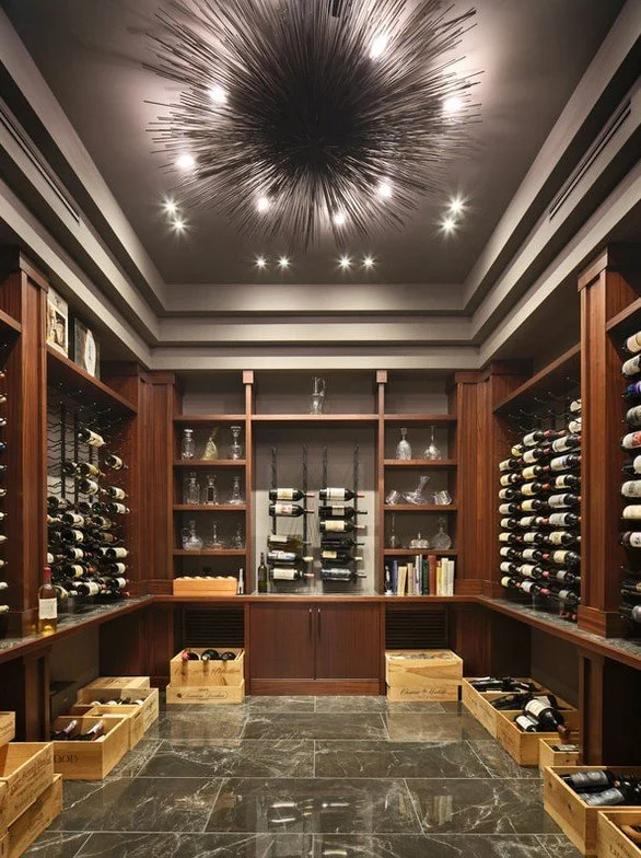 A luxurious wine cellar with dark wood cabinets, wine racks filled with bottles, glassware, books, and wooden crates on a marble floor, illuminated by a modern black starburst chandelier on the ceiling.