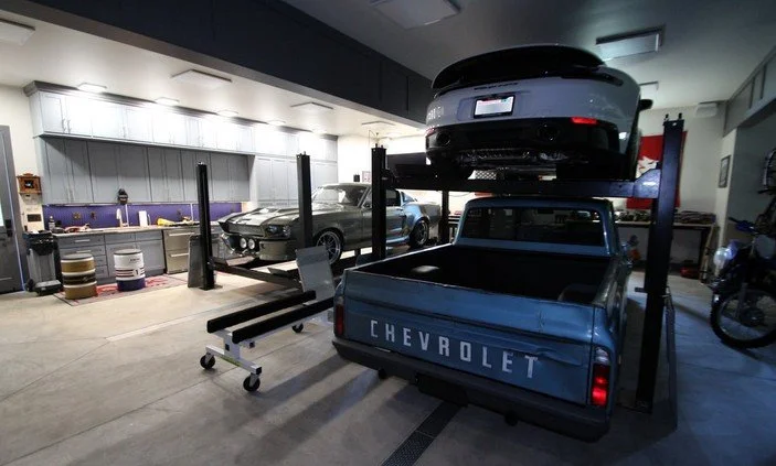 An indoor garage with vintage cars and a Chevrolet pickup truck, with one car elevated on a lift.