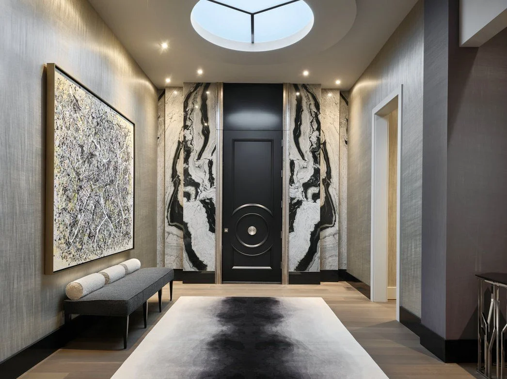Modern entryway featuring a black door with geometric design, black and white marble accents, a large abstract painting, a gray bench with pillows, and a circular skylight ceiling.