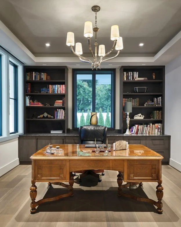 A modern office with a wooden desk, black leather chair, bookshelves filled with books, a window showing greenery outside, and a chandelier hanging from the ceiling.