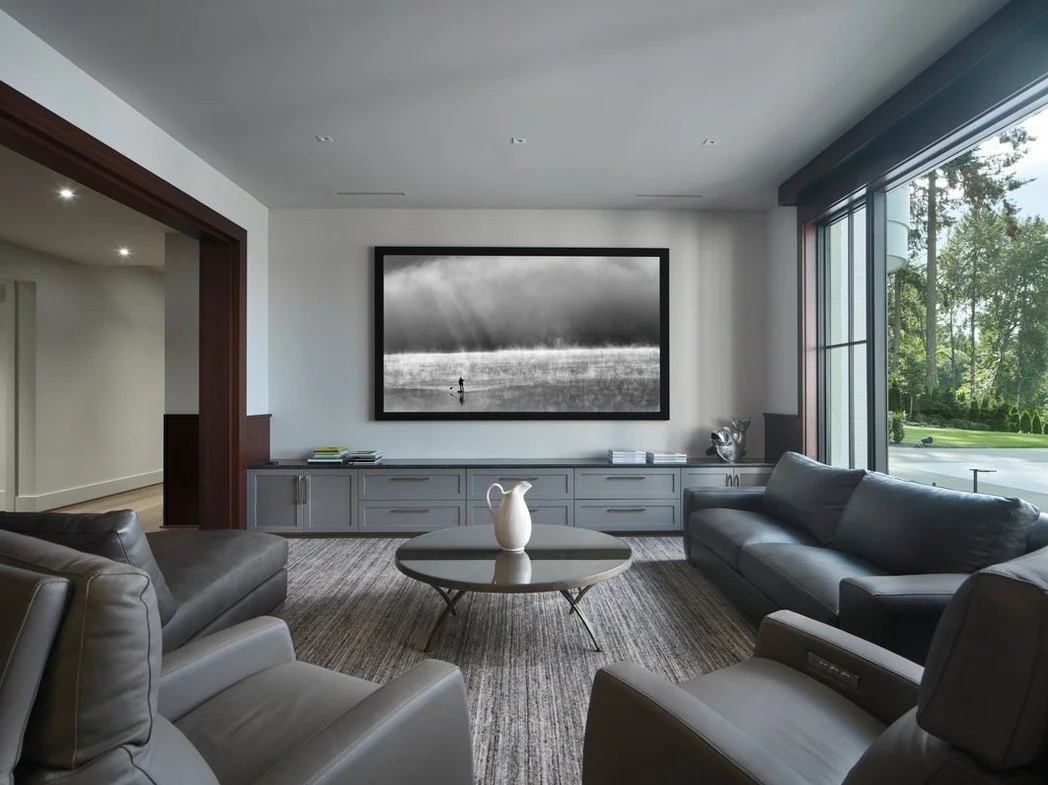 Modern living room with large glass windows, gray sofas, a round coffee table with a white pitcher, and a framed black-and-white landscape photograph on the wall.