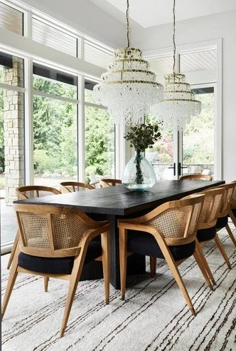 Dining room with a dark wood table, six wicker chairs with cushions, large glass vase with flowers, two ornate chandeliers, large windows with green trees outside, and a striped rug.