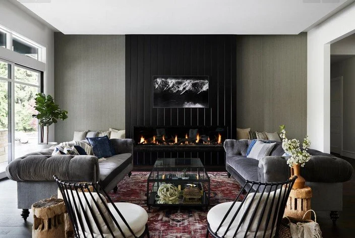 Modern living room with two gray velvet sofas, a black fireplace, a glass coffee table, and a patterned area rug. Large windows on the left side letting in natural light, with a potted plant near the window. Wall art above the fireplace, decorative c