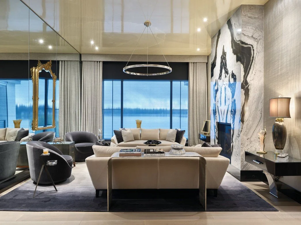 Luxury living room with beige sofa, black armchairs, large window overlooking water, marble fireplace, gold mirror, decorative lamps, and modern art pieces.