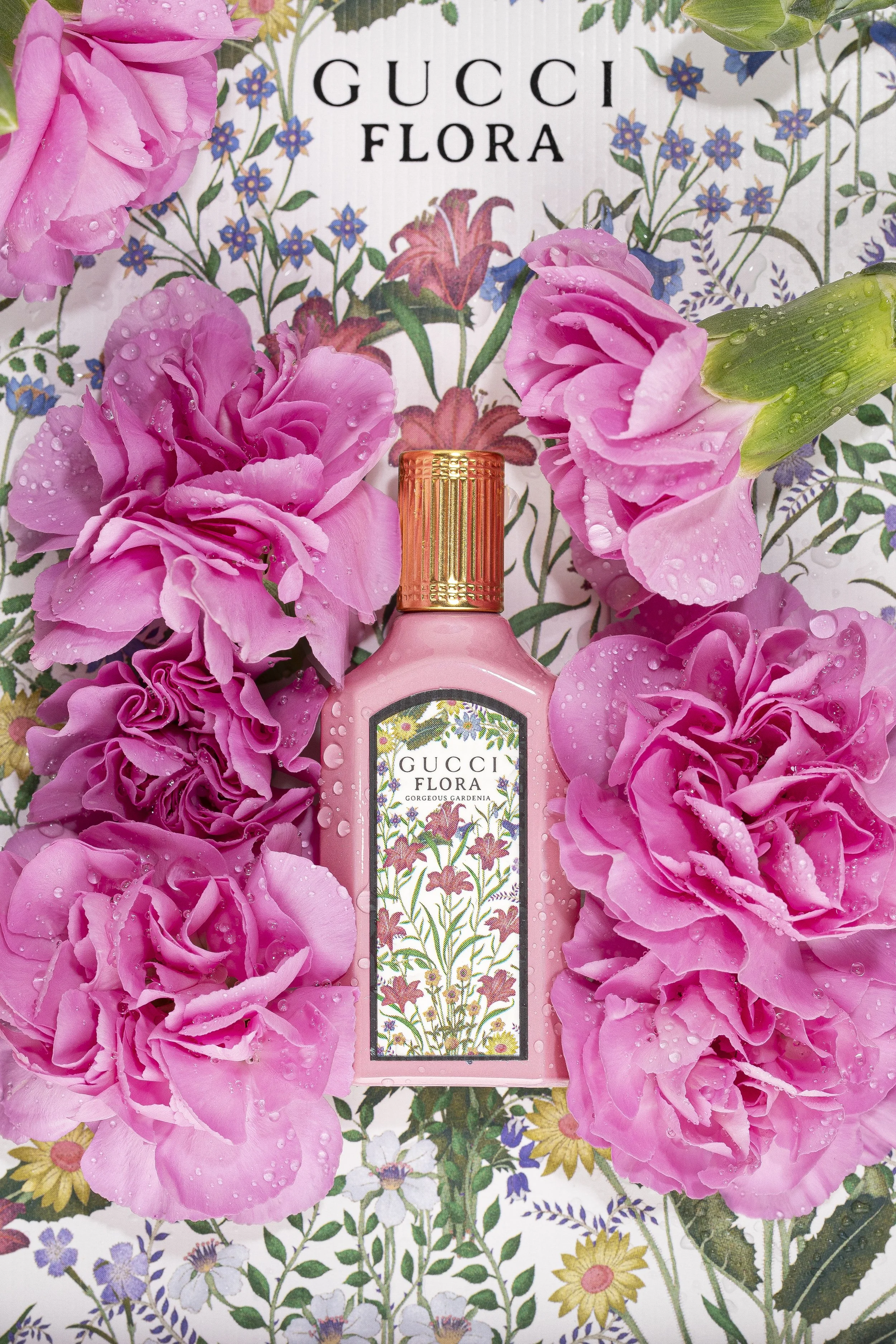 A bottle of Gucci Flora perfume surrounded by pink flowers and green leaves, with a floral background that says 'GUCCI FLORA' and 'GORGEOUS GARDENIA'.