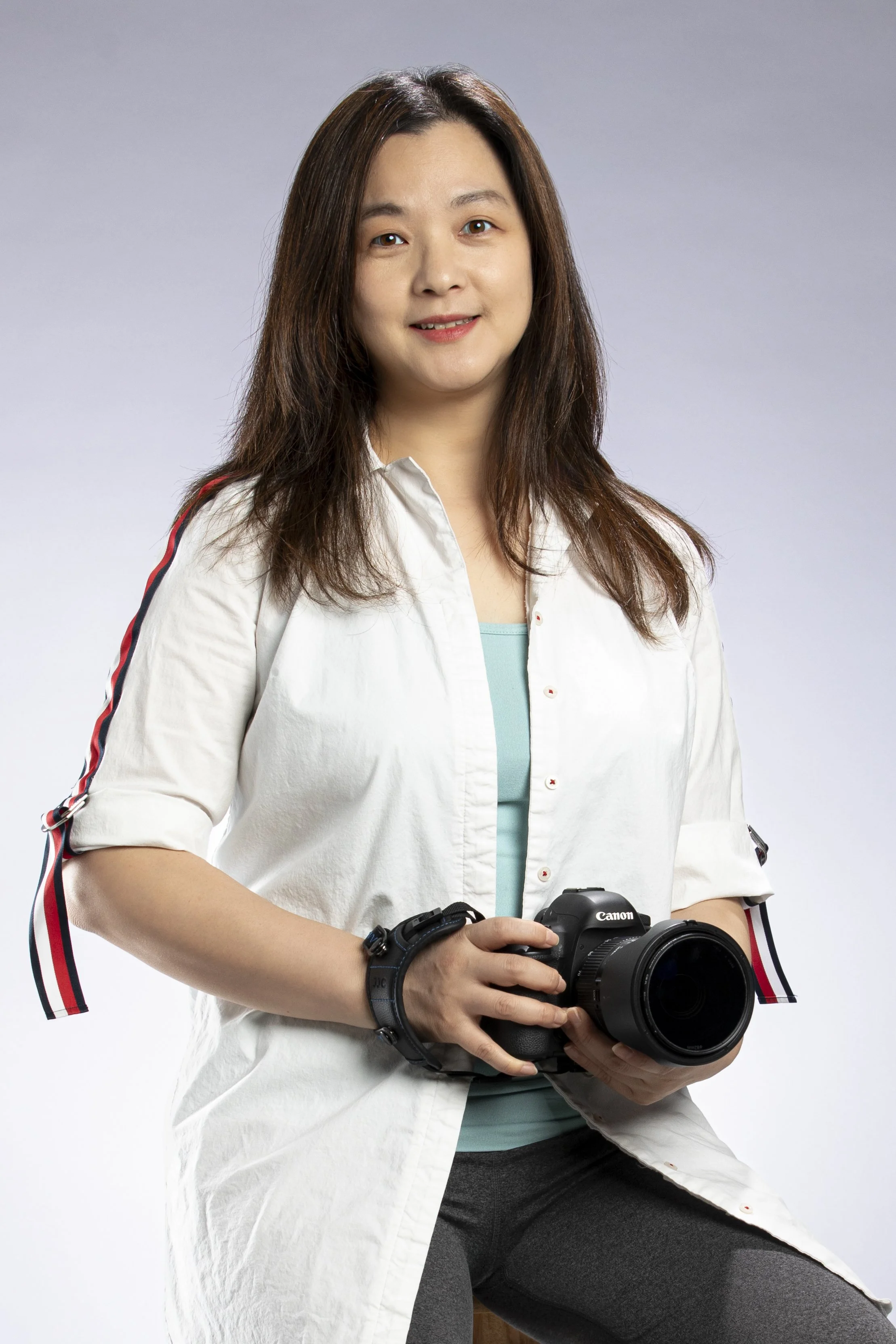 A woman with shoulder-length brown hair wearing a white shirt with red, white, and blue stripes on the sleeves, holding a Canon camera, standing against a plain background.