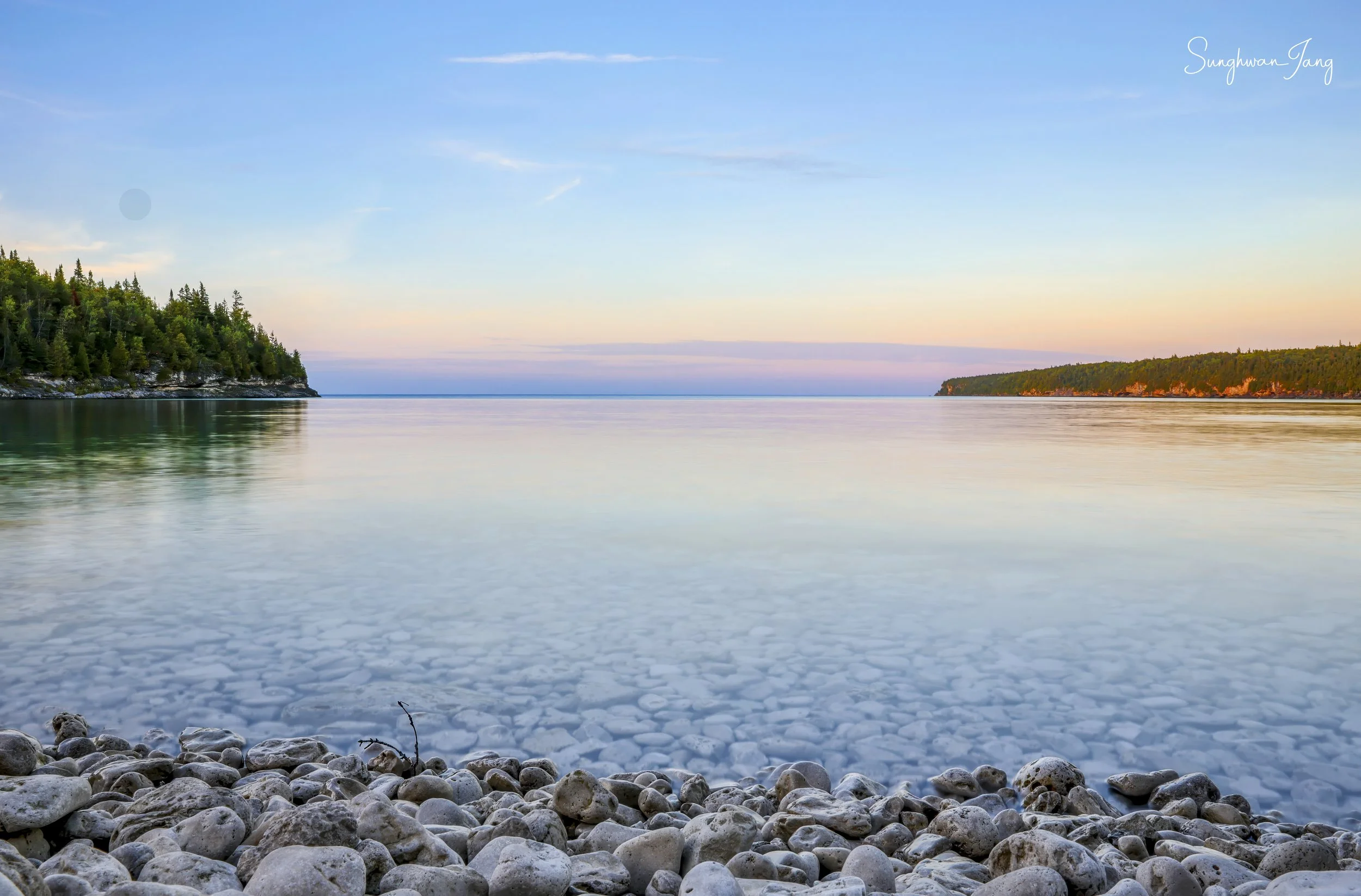 Calm water with ice and rocks in the foreground, green trees on the left and right shoreline, clear sky with soft sunset colors in the background.
