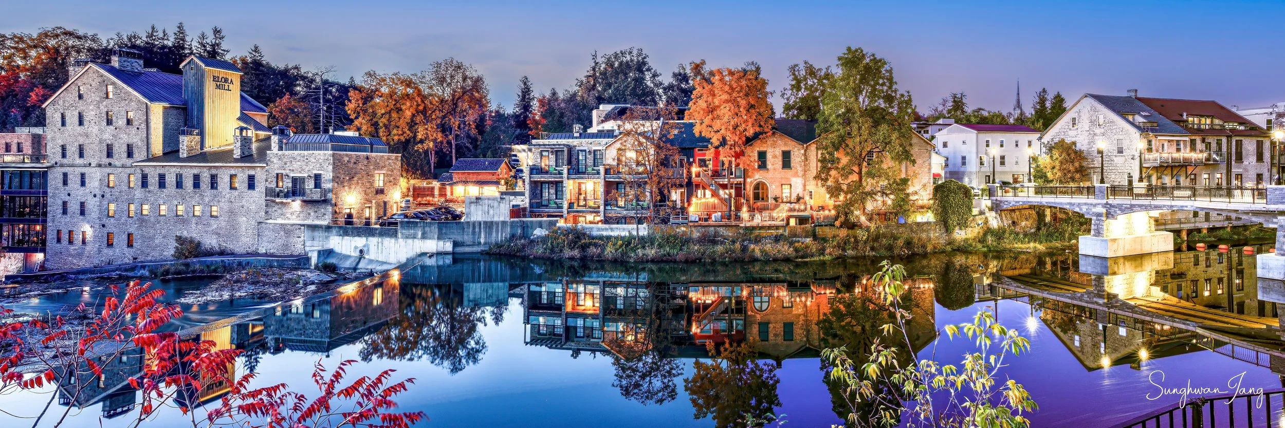 A twilight view of a town with a river reflecting the illuminated buildings and trees, featuring a mix of modern and historic architecture, with colorful autumn foliage.