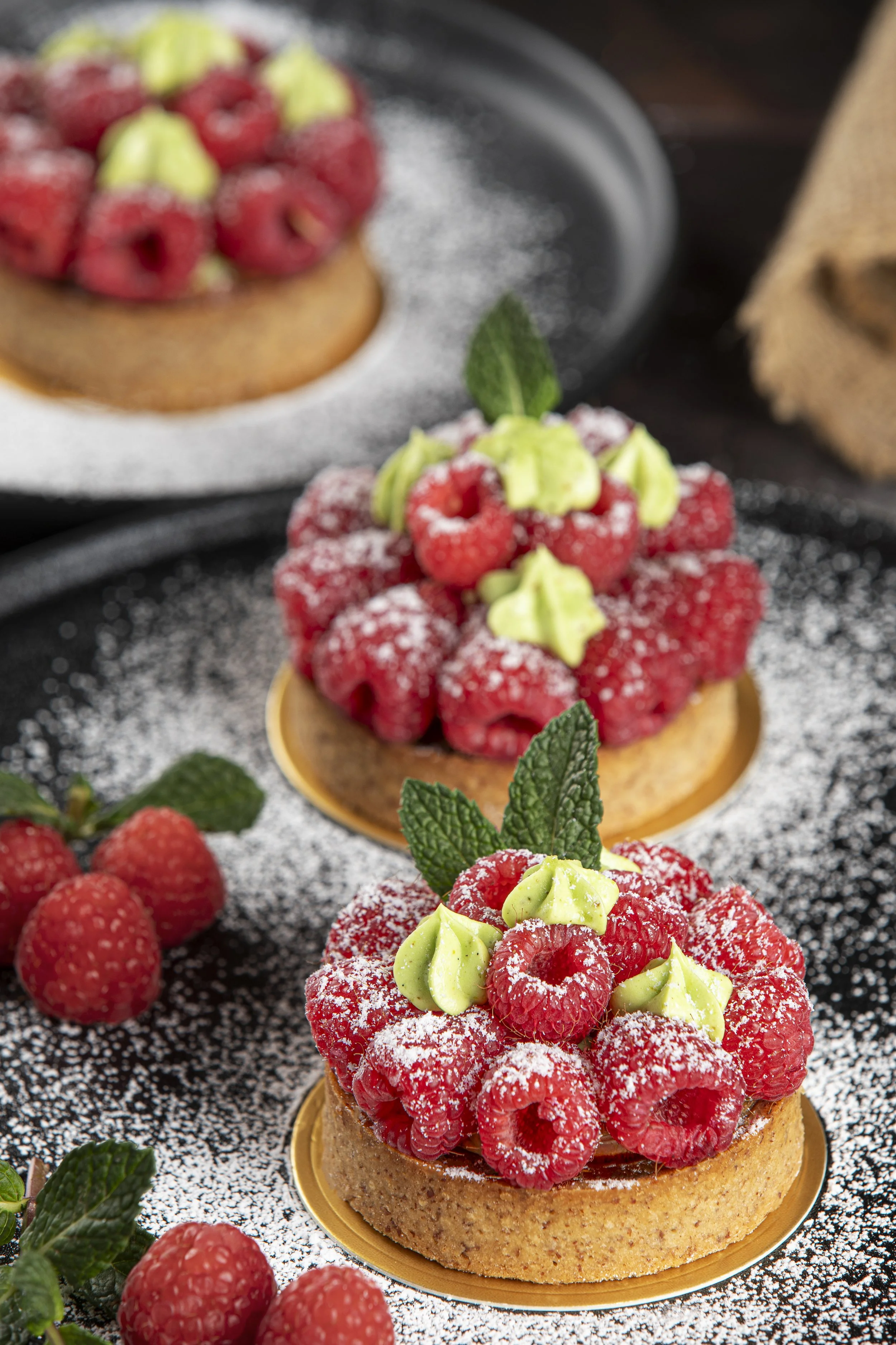 Two raspberry tarts topped with raspberries, green cream, mint leaves, and powdered sugar, on a black surface with extra raspberries and mint leaves around.