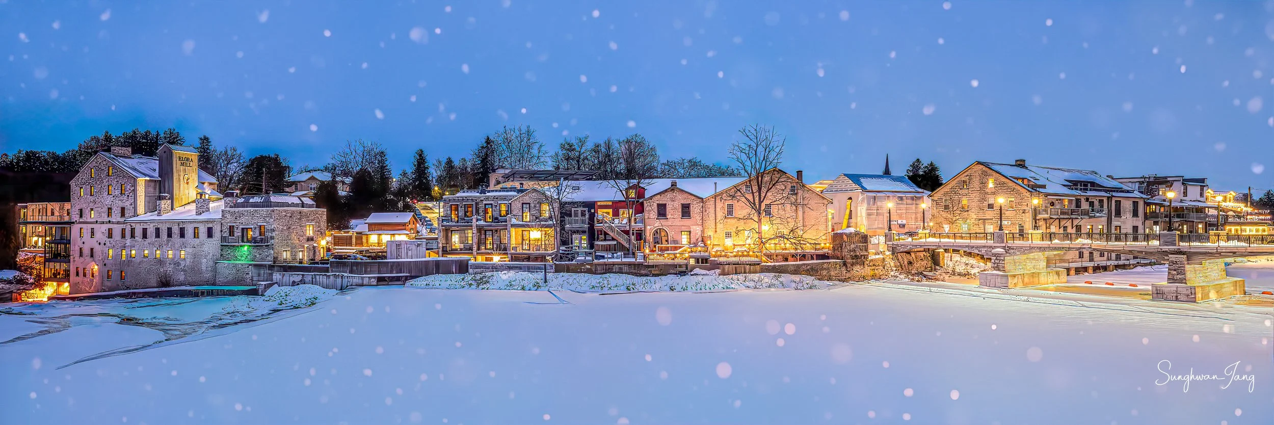 Snow-covered town with illuminated buildings and frozen river at night, with falling snow.