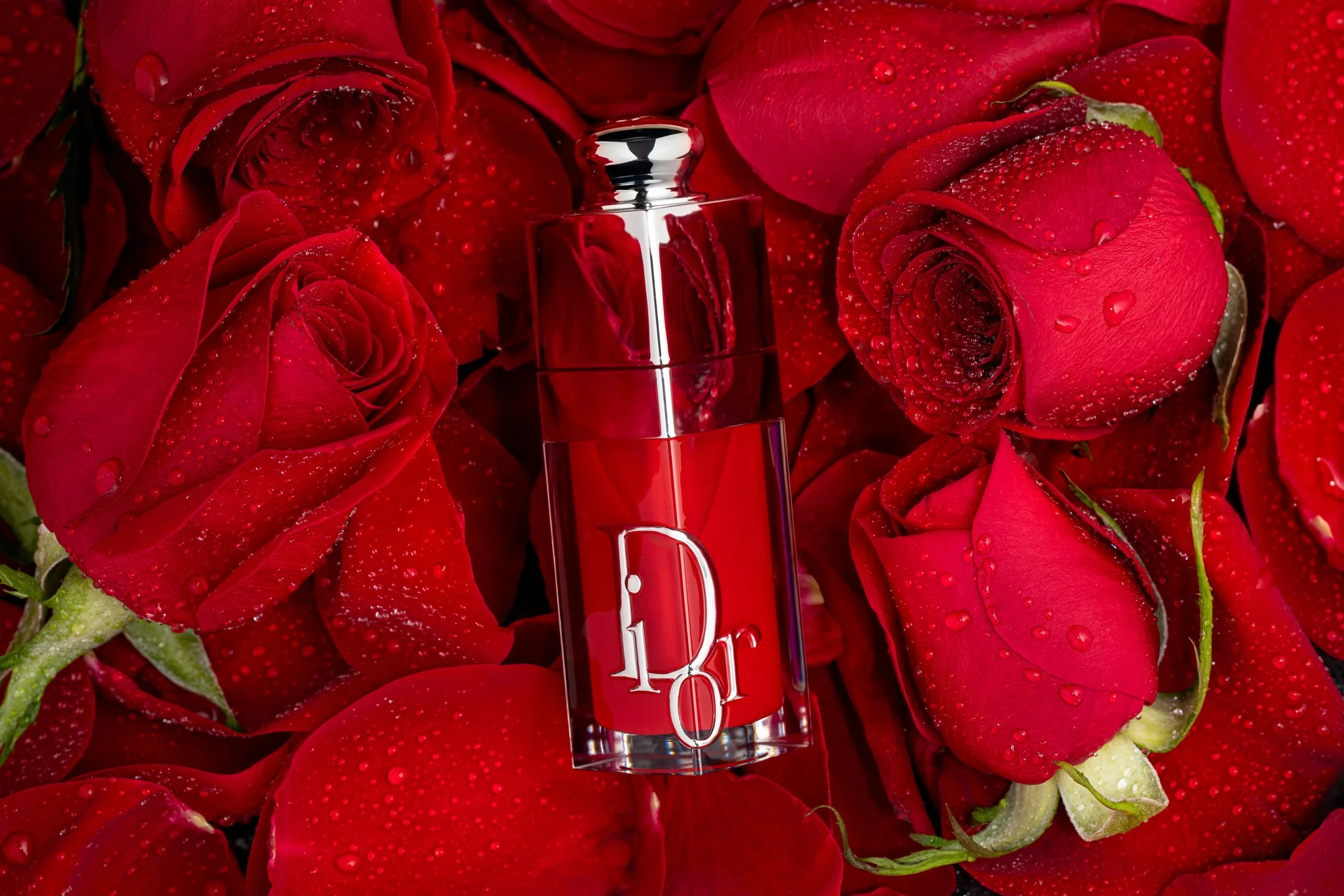 A bottle of Dior perfume surrounded by red roses with water droplets.