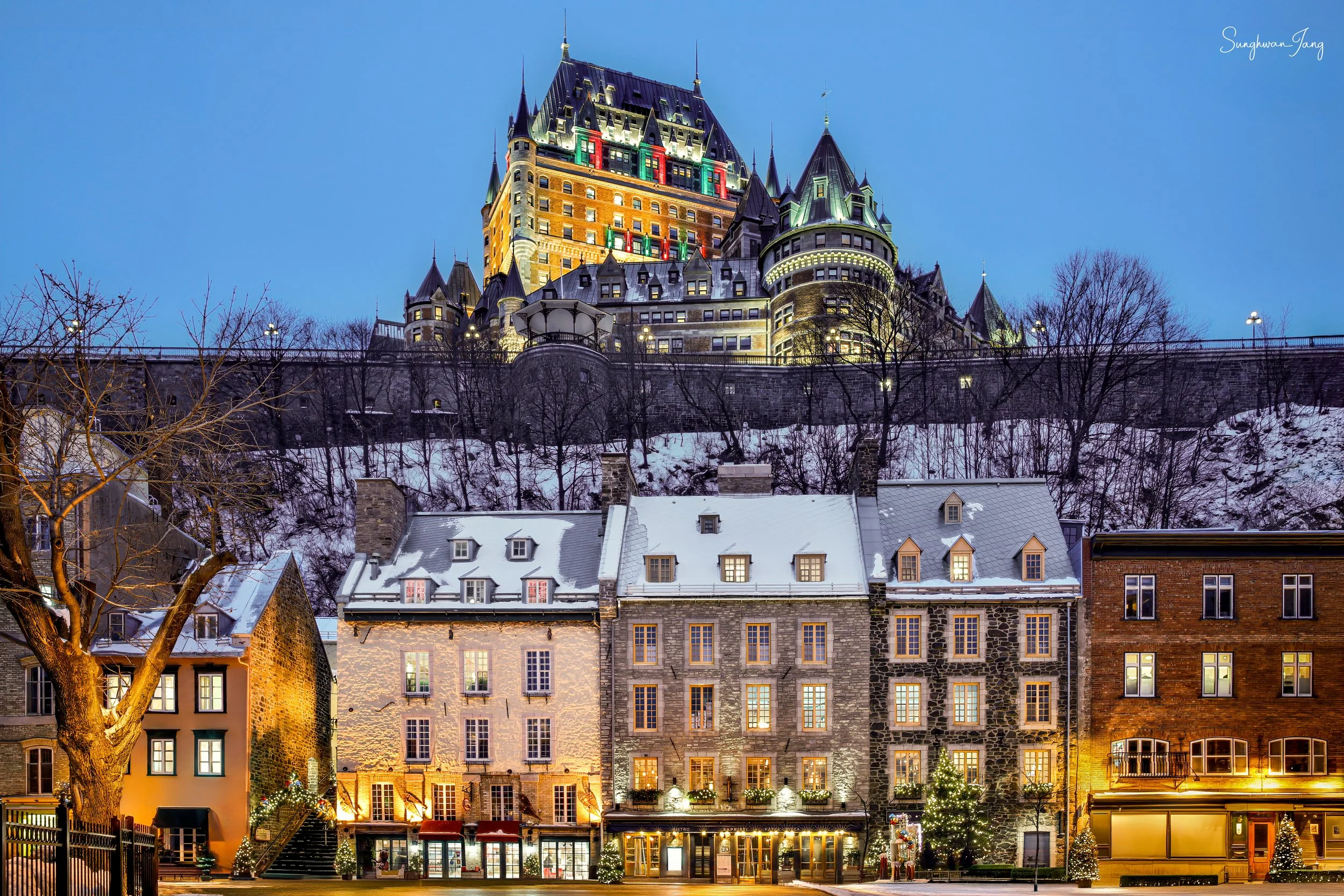 Night view of a castle on a hill illuminated with colorful lights, overlooking a row of historic buildings decorated with Christmas lights in a snowy setting.