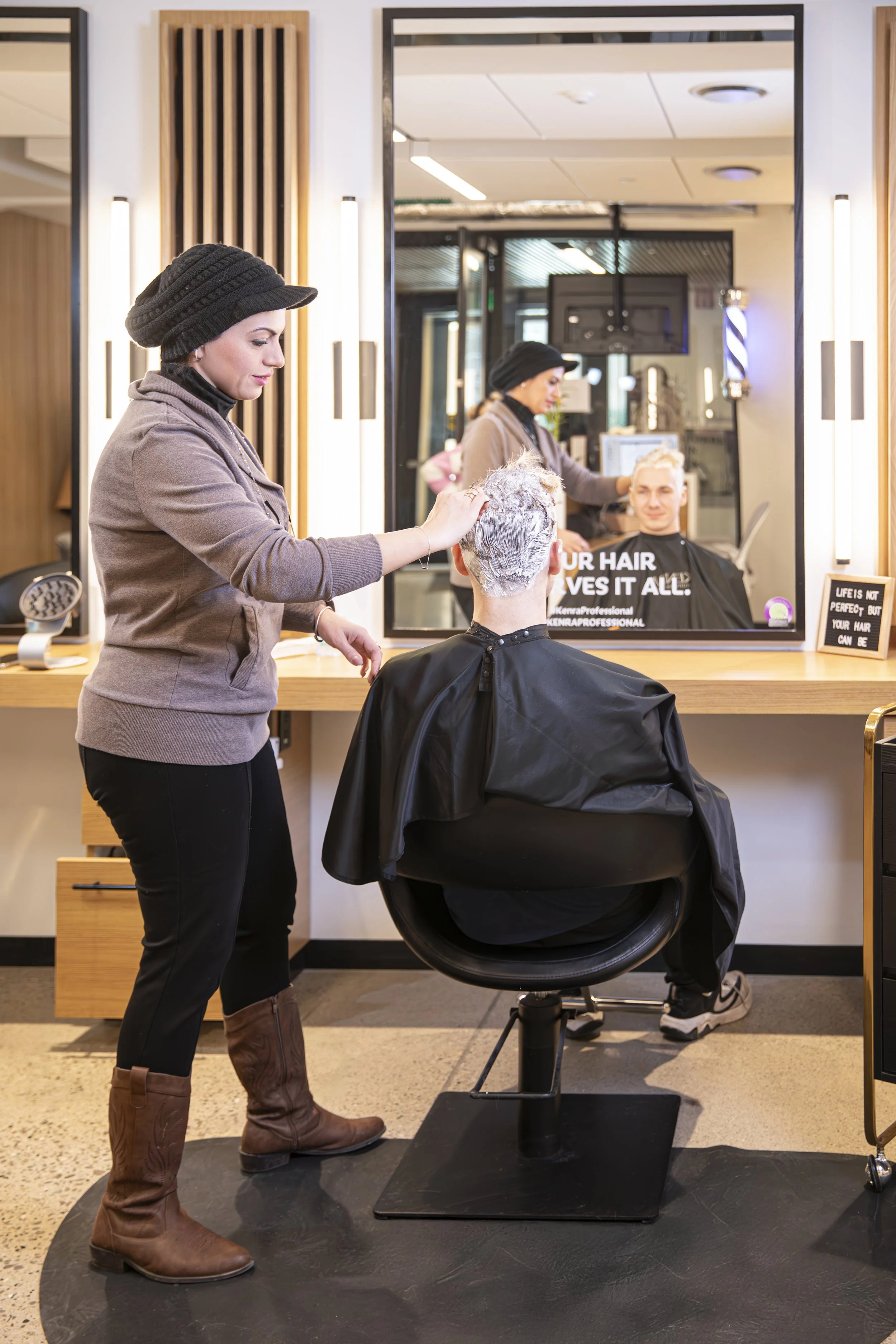 A hairstylist applying hair dye to a customer's hair in a salon. The customer is seated in a black salon chair, wearing a black cape, and has their hair covered in a white foam or bleach. There is a large mirror in front of the customer, reflecting the stylist working on their hair. The salon has warm lighting, wood accents, and a sign on the counter that reads, "Life is not perfect but your hair can be."