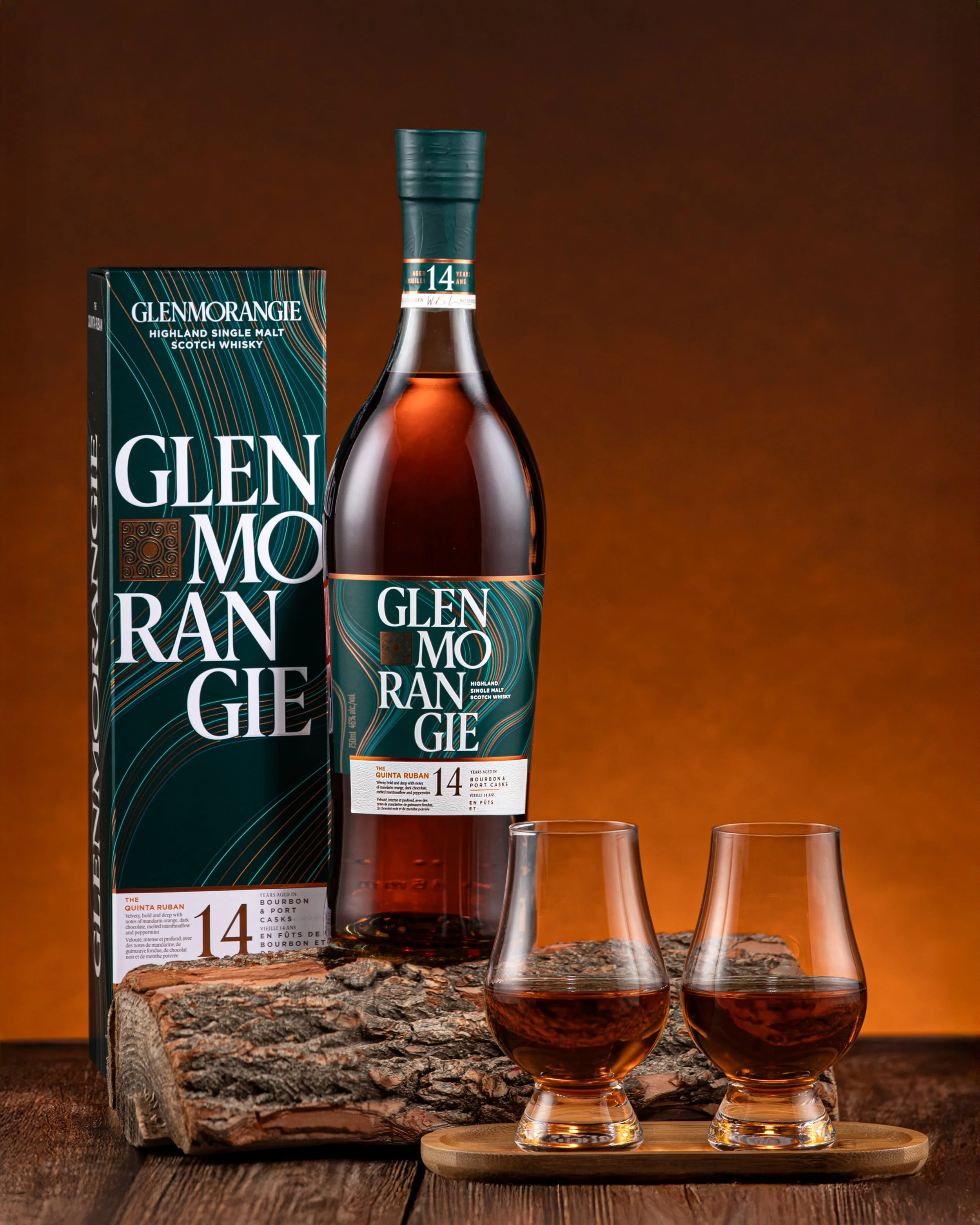 A bottle of Glen Morangie Highland Single Malt Scotch Whisky, with its box, placed on a log, alongside two whisky glasses filled with whisky, against a brown background.
