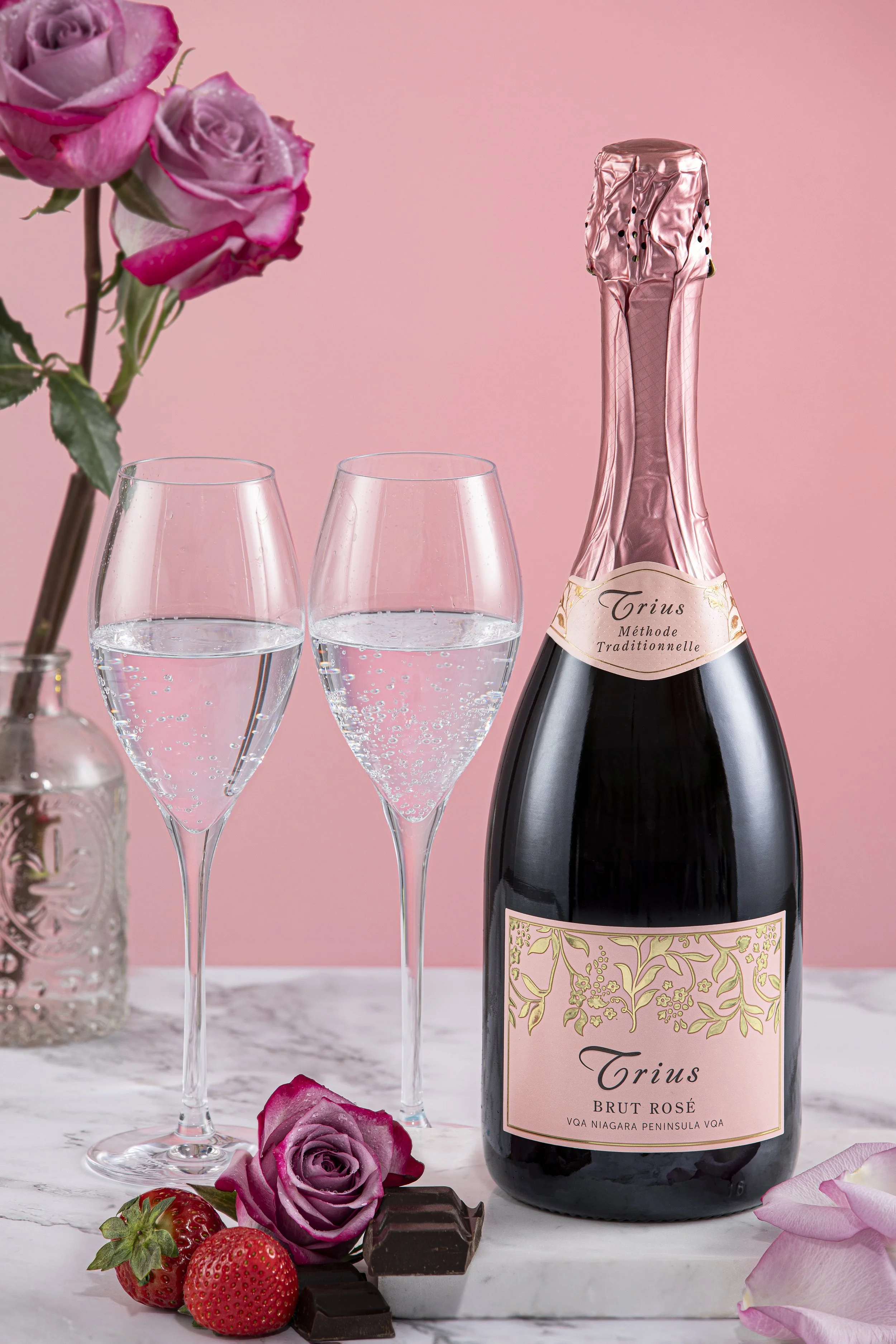 A bottle of pink bubbly sparkling wine, two filled champagne glasses, pink roses, strawberries, and chocolates on a marble surface, with a pink background.