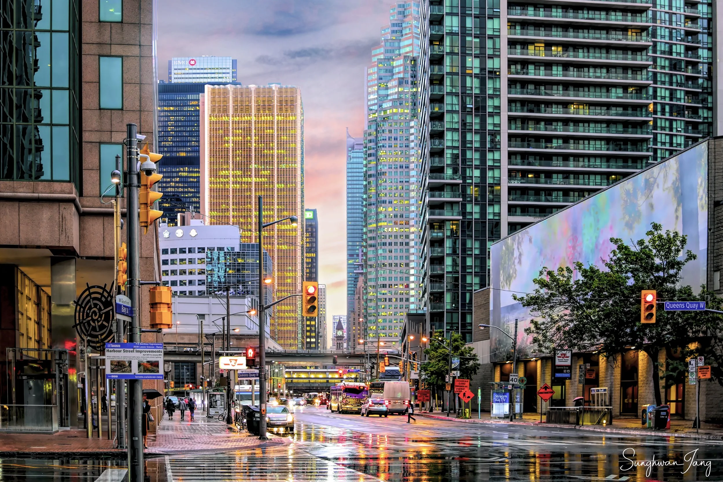 Rainy city street scene at dusk with tall modern buildings, traffic lights, cars, buses, pedestrians, and wet pavement reflecting lights, in downtown