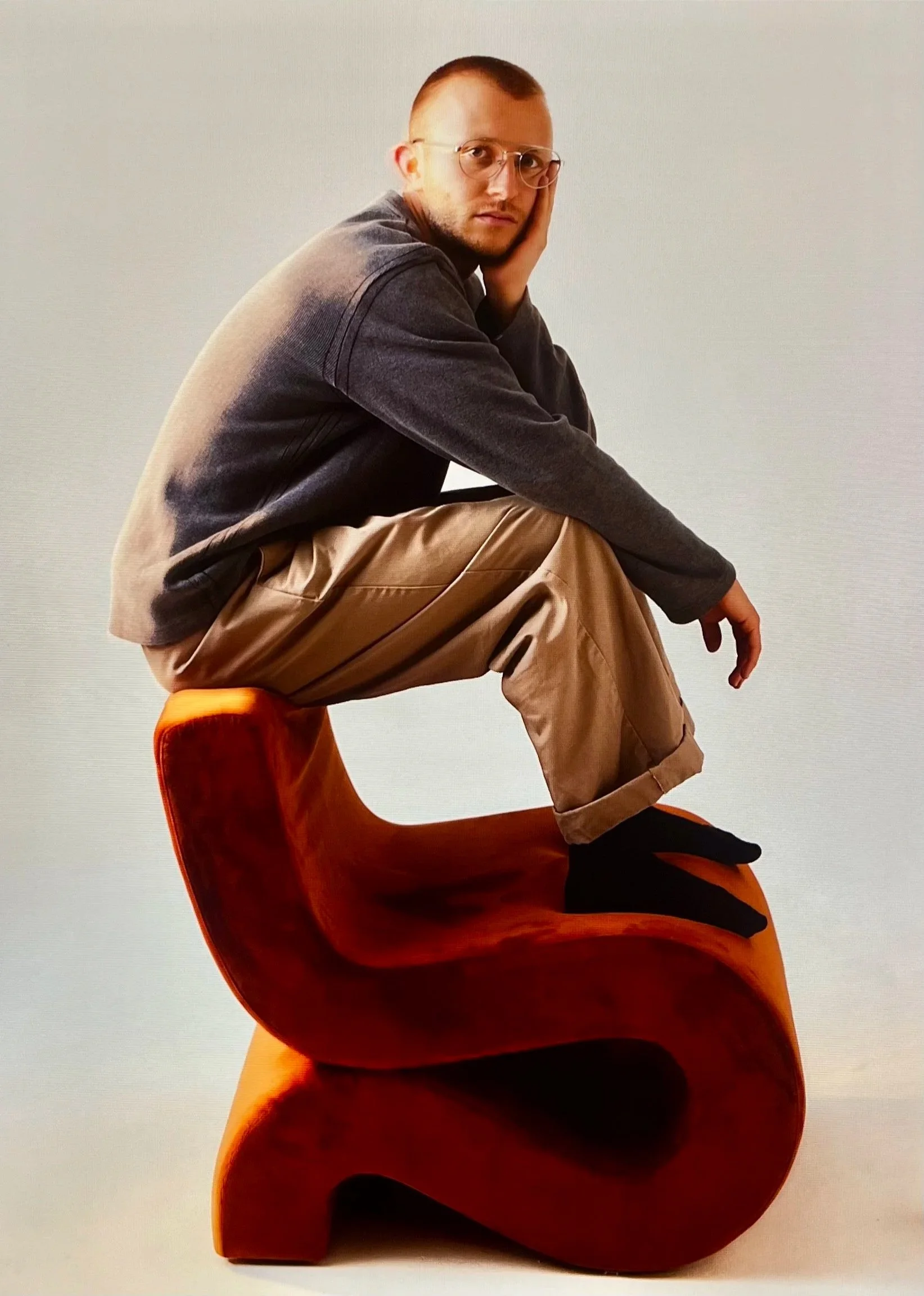 A man wearing glasses, a gray sweatshirt, and khaki pants sits on a large, abstract, orange sculpture with a curved, wave-like shape, against a plain background.