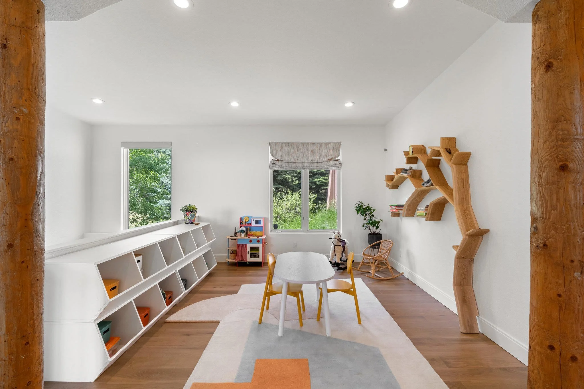 Child's playroom with white walls and wooden floor, featuring a round table with four yellow chairs, a white angled bookshelf, a toy kitchen, a rocking chair, a plush rocking horse, a potted plant, and wooden wall shelves, with large windows showing 