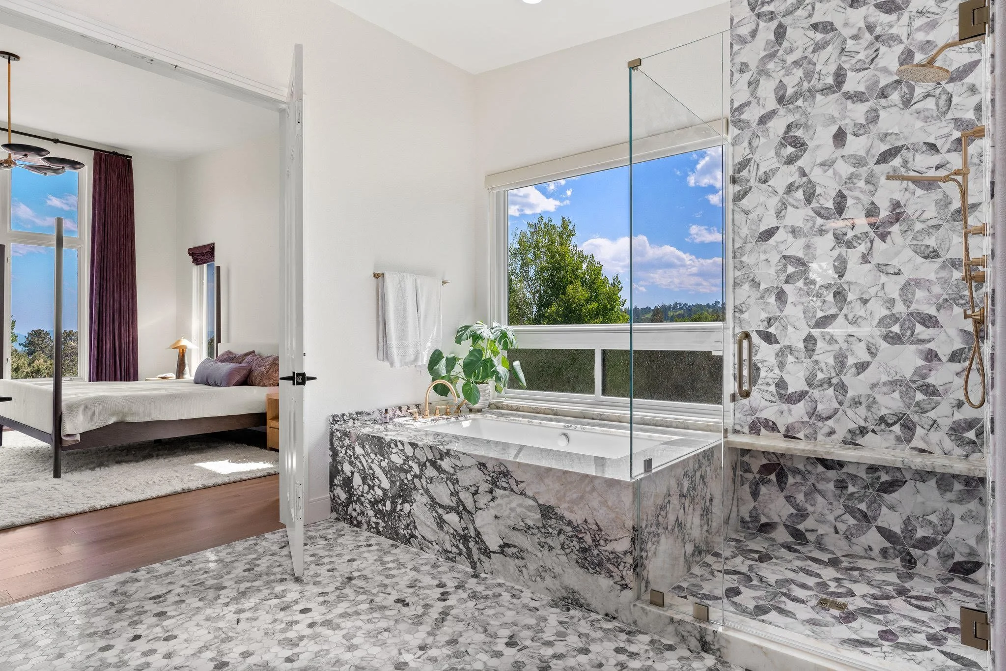 Modern bathroom with marble bathtub and walk-in shower, large window showing blue sky and trees, and bedroom with bed and purple curtains visible through doorway