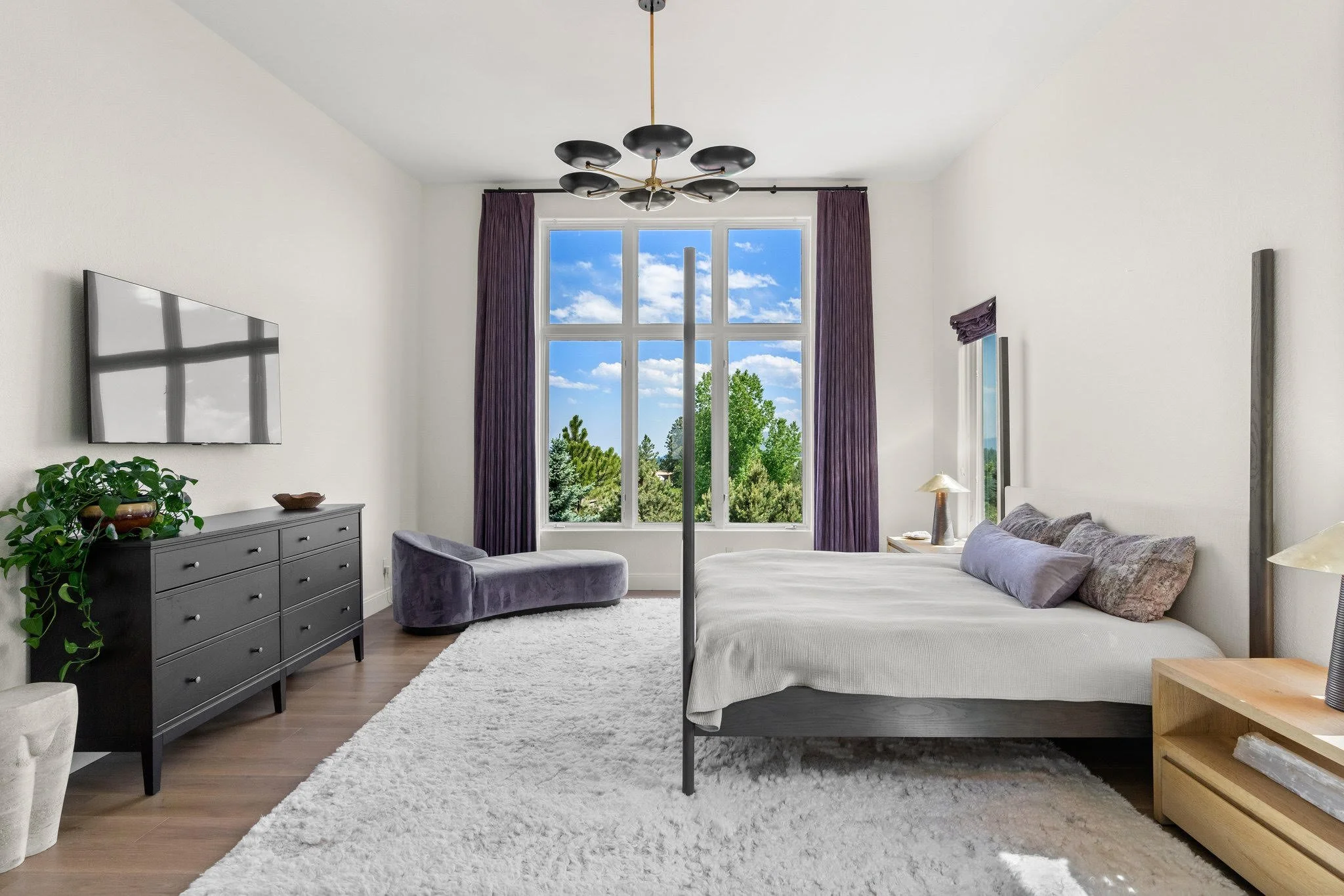 Modern bedroom with a large window showing trees and a blue sky, a bed with gray pillows, a black dresser with a green plant, purple curtains, a gray chaise lounge, a white rug, and a ceiling light fixture.