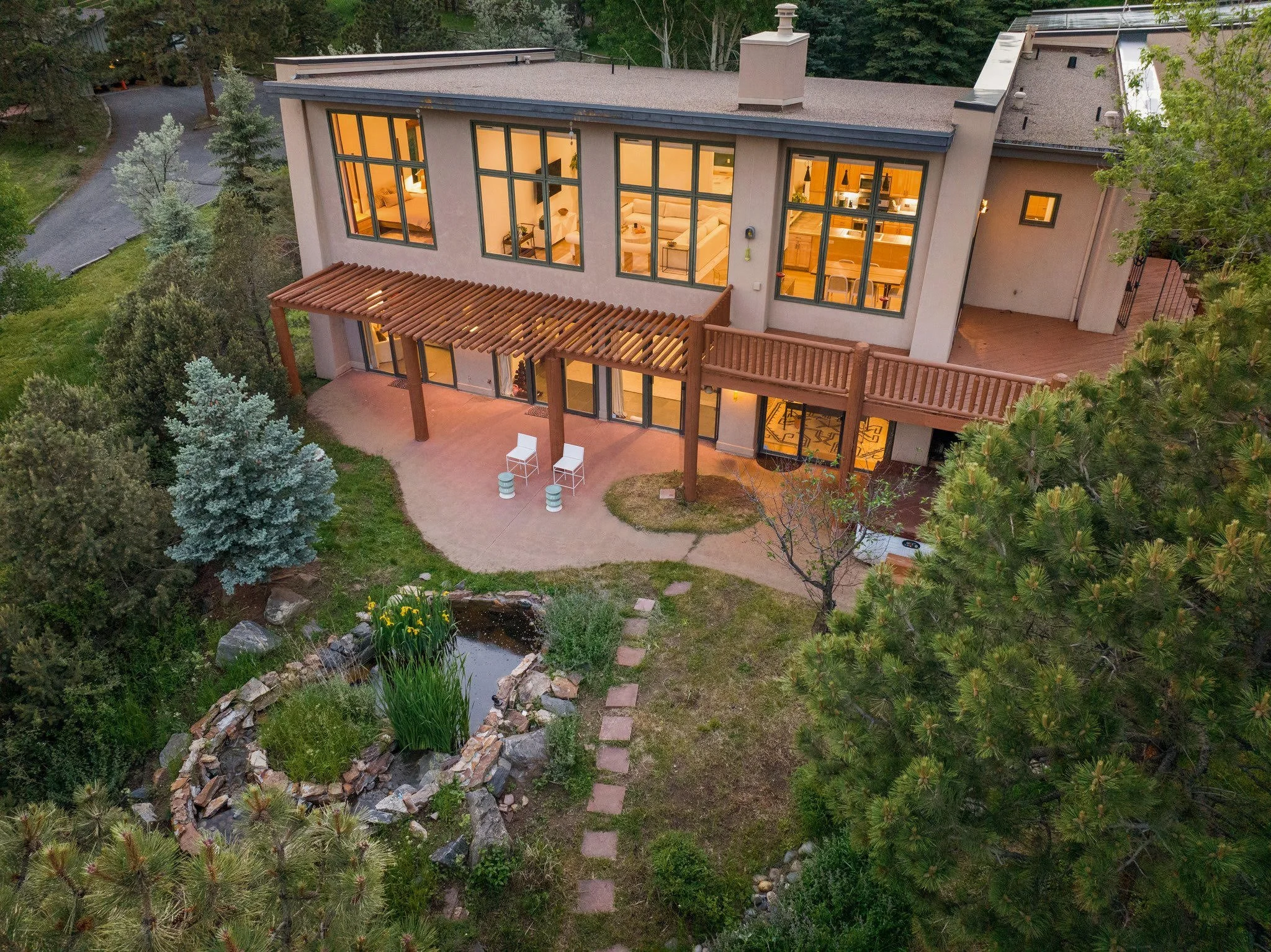 A modern two-story house with large windows, a wooden balcony, and a landscaped backyard with trees, a small pond, and a stone pathway.