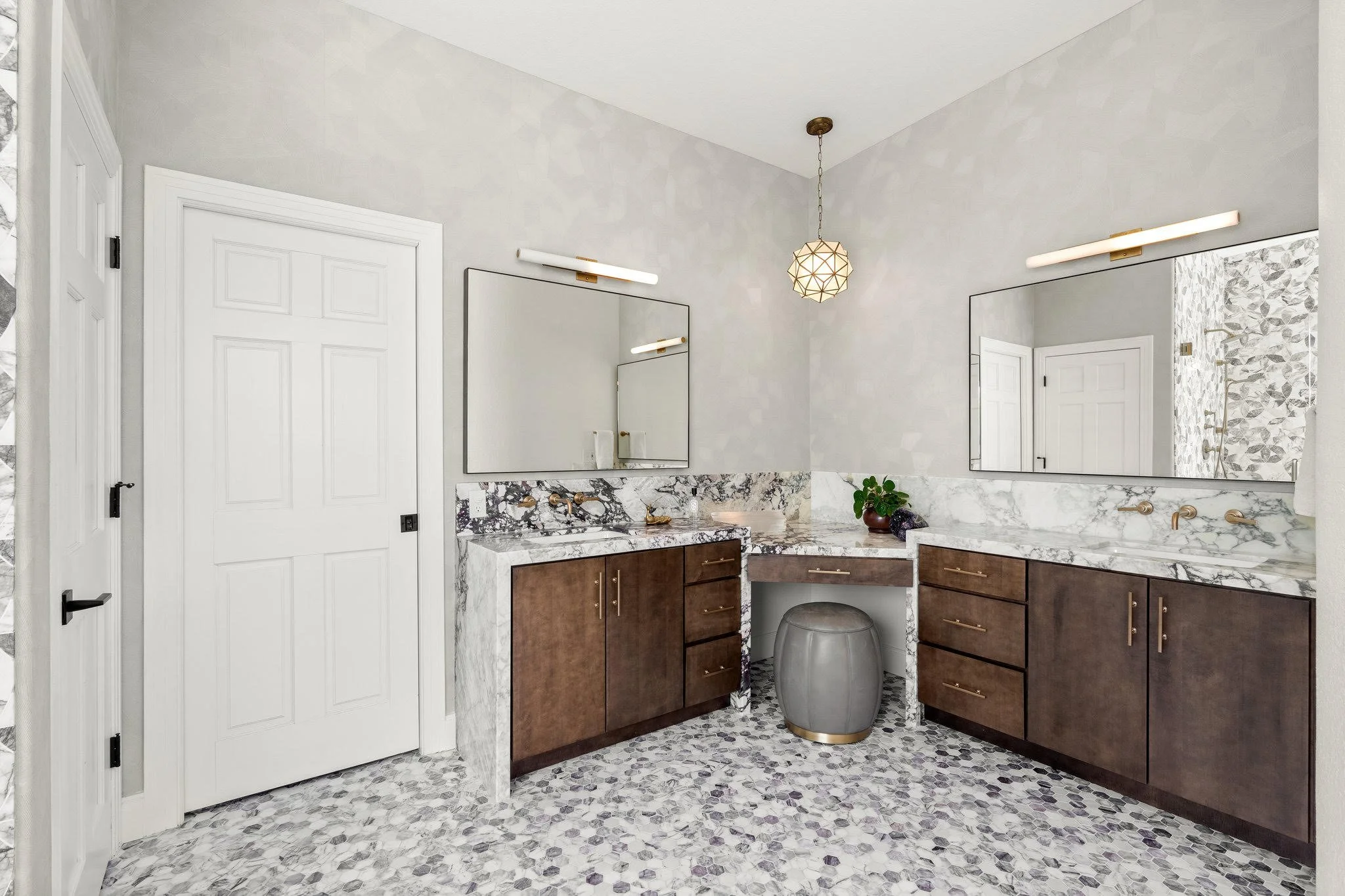 Modern bathroom vanity with marble countertops, two large mirrors, and brown cabinetry, with a pebble tile floor, a small grey stool, and a lamp hanging from the ceiling.