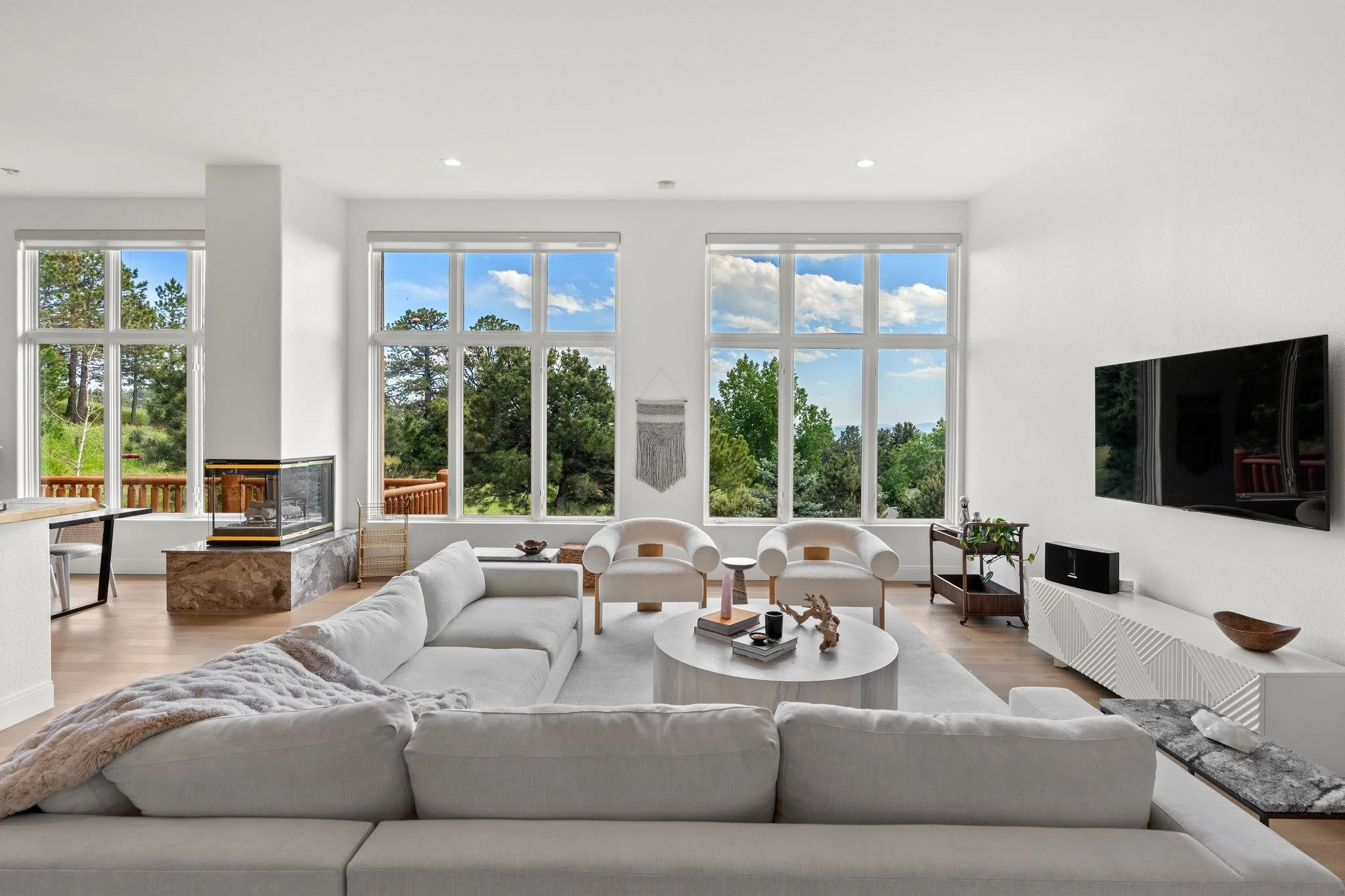Modern living room with large windows, white sofa, two white armchairs, a wall-mounted TV, and decorative items