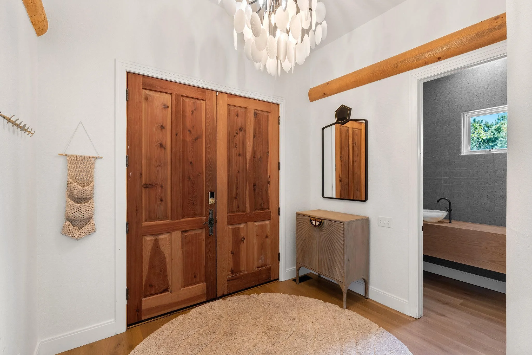 Entryway with wooden double doors, white walls, a decorative light fixture, a mirror, a small wooden cabinet, a beige rug, and a view into a bathroom with a small window and sink.