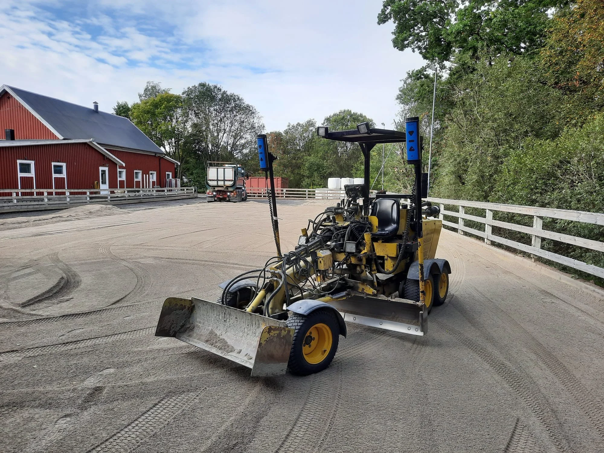 A yellow compact construction vehicle working on a gravel surface near a white wooden fence on a farm or rural area.