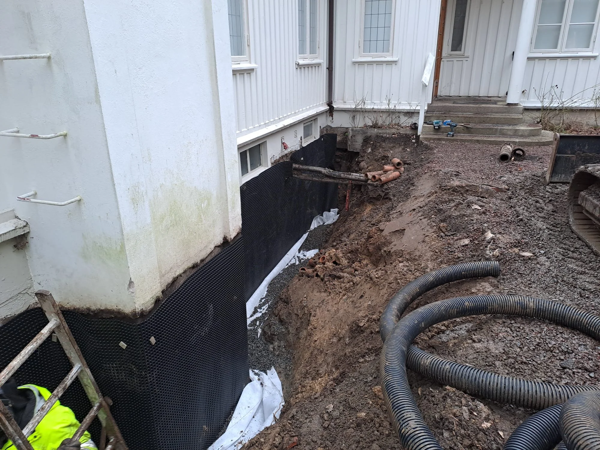 Construction site with exposed ground, black waterproof barrier installed along the foundation of a house, pipes, and construction tools.