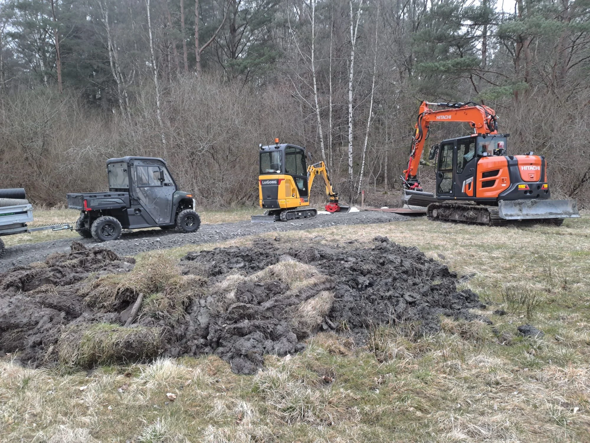 Construction site with a small orange excavator, a gray utility terrain vehicle, and a larger orange excavator with a forest background.