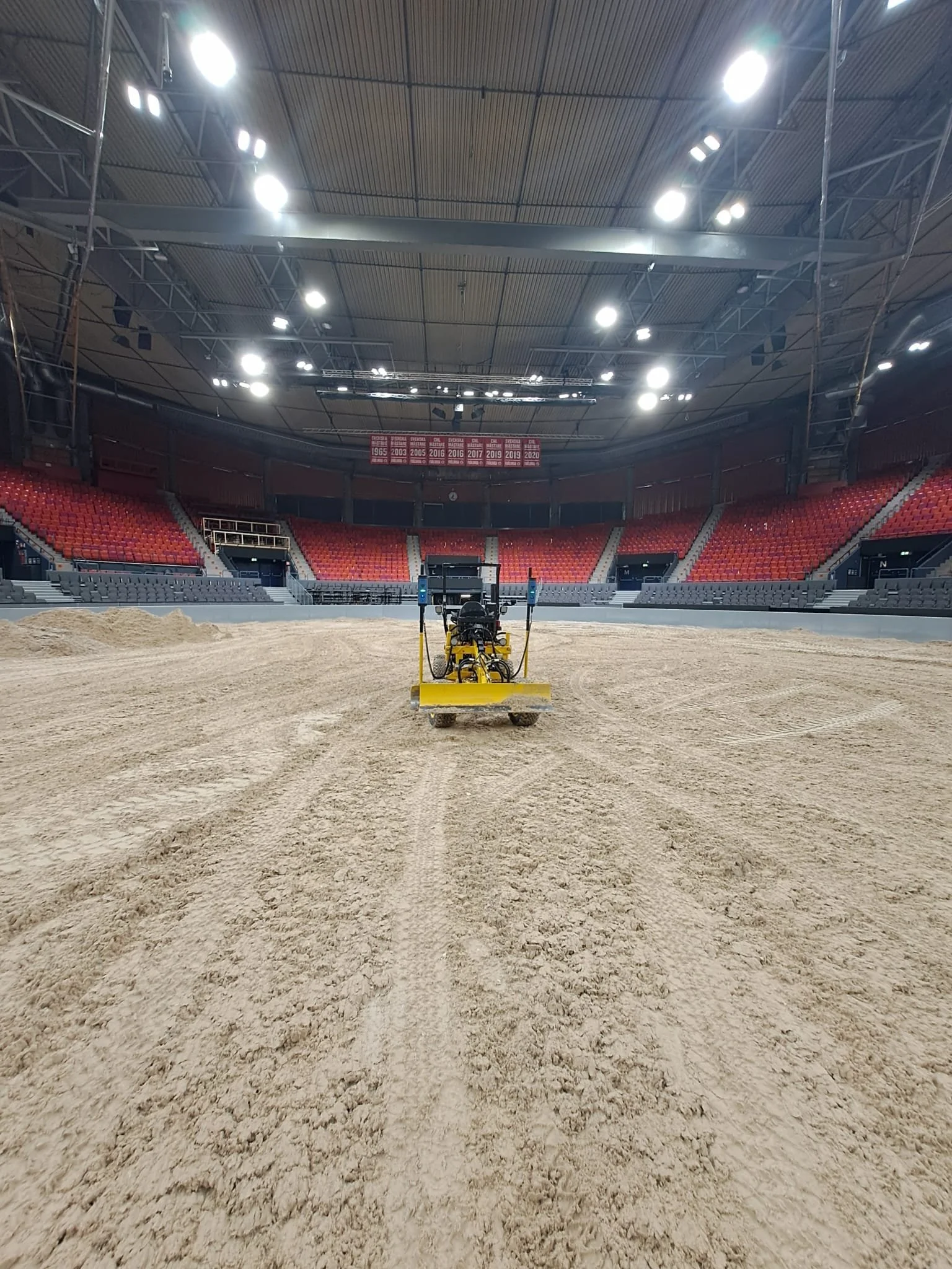 Indoor arena with sand floor and construction equipment in the center, empty red and black seating surrounding the space, and bright overhead lighting.