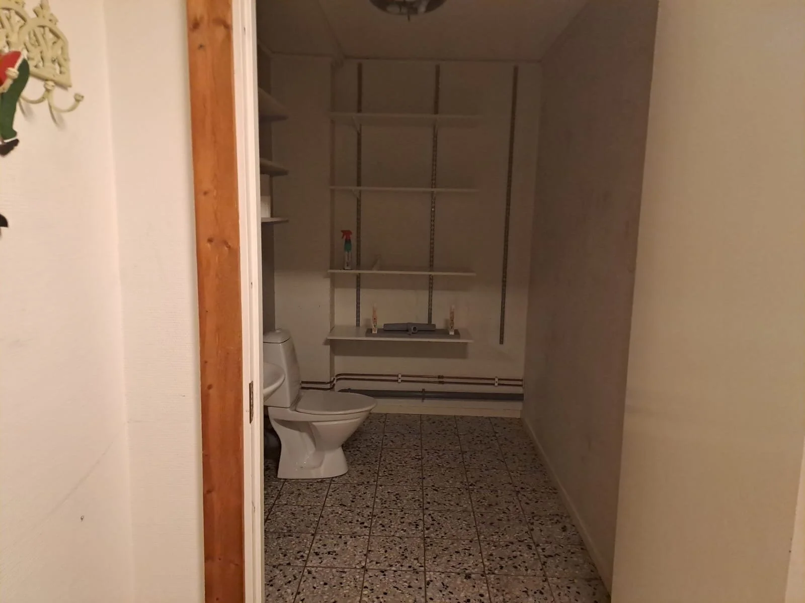 Ett tomt badrum med en toalett, tomma vita hyllor, ett sprayflaska och några små föremål på en hylla.