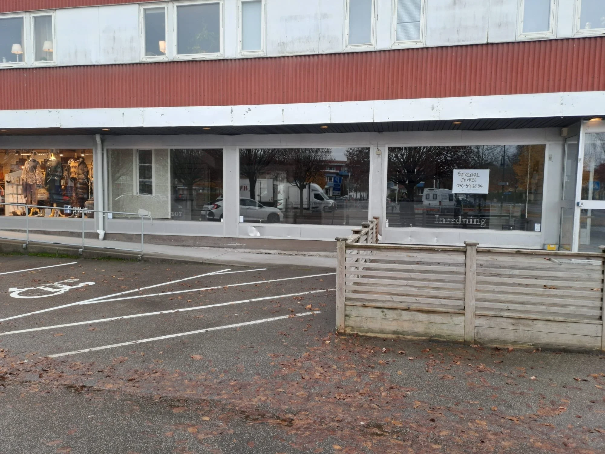 Bild på en tom parkeringsplats med handikappparkeringmarkerad plats framför en butikslokal med stora fönster. Utanför butiken finns en liten trästaket och ett skylt som visar att det är en butik som hyrs ut.