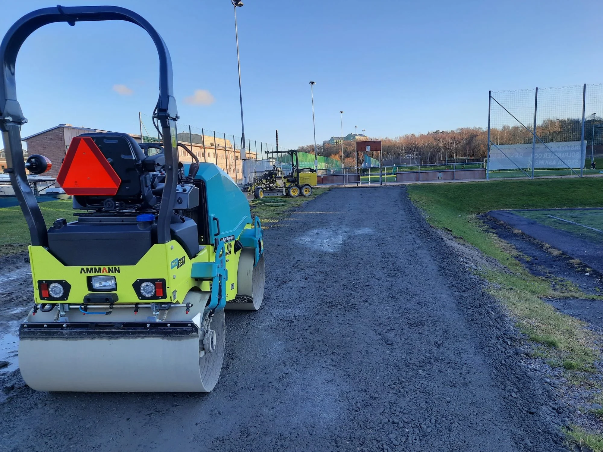 A construction road roller compacting gravel on a pathway at a sports field, with additional construction equipment and a fenced sports field in the background.