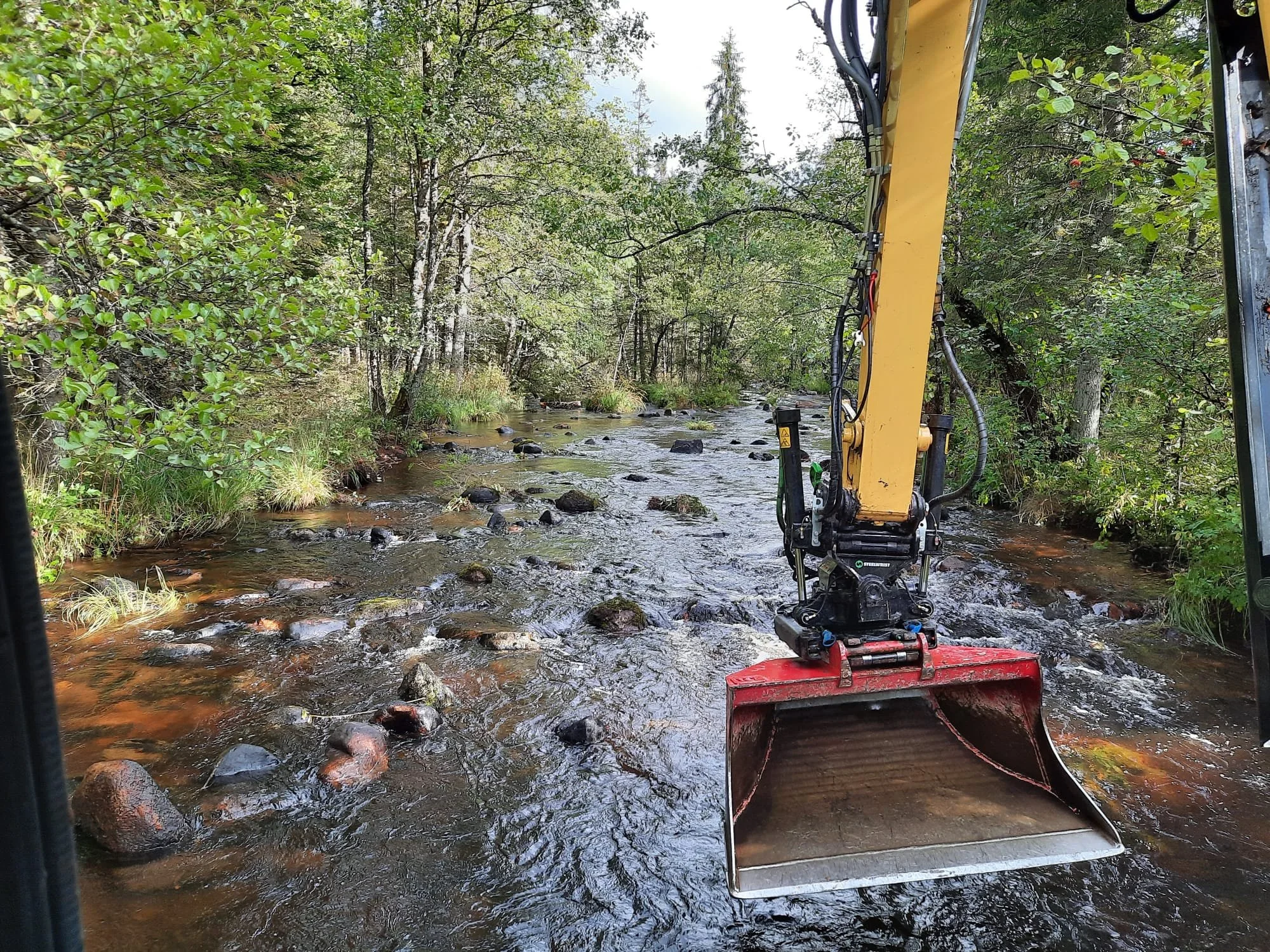 View from a moving vehicle on a shallow river or creek with rocks and surrounding trees, as a yellow excavator with a red bucket is operating in the water.