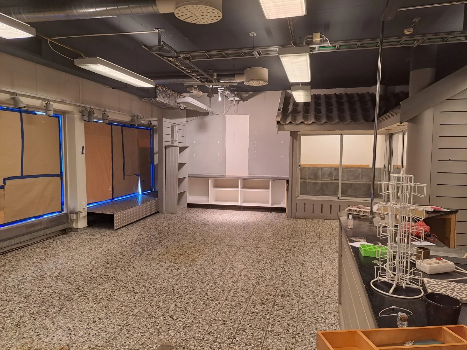 Tomt café eller butikslokal med låsta fönster och tom diskbänk, dekorationsställ, förvaringsskåp, hyllor och takbelysning.