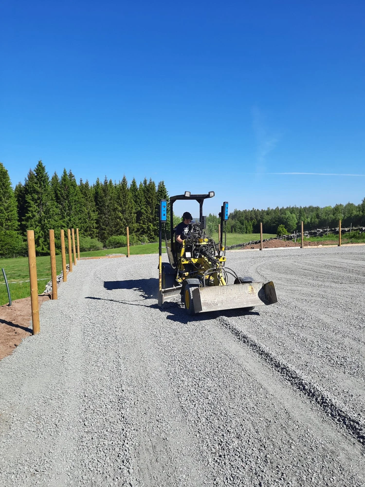 En person som kör en hyvel på en grusplan med gröna träd i bakgrunden under en klar blå himmel.