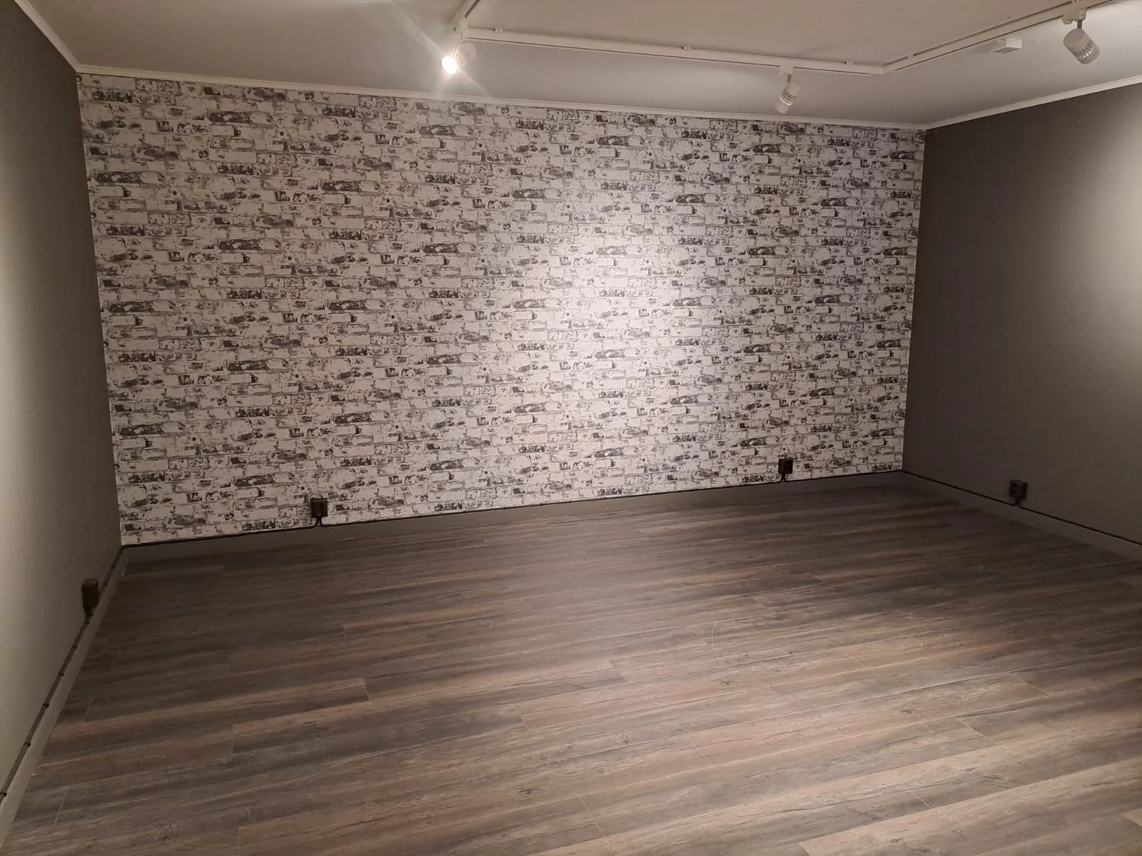 Ett tomt rum med träfärgad golv och väggar, en med vit tegelvägg och två i mörkare färg, med spottar i taket.