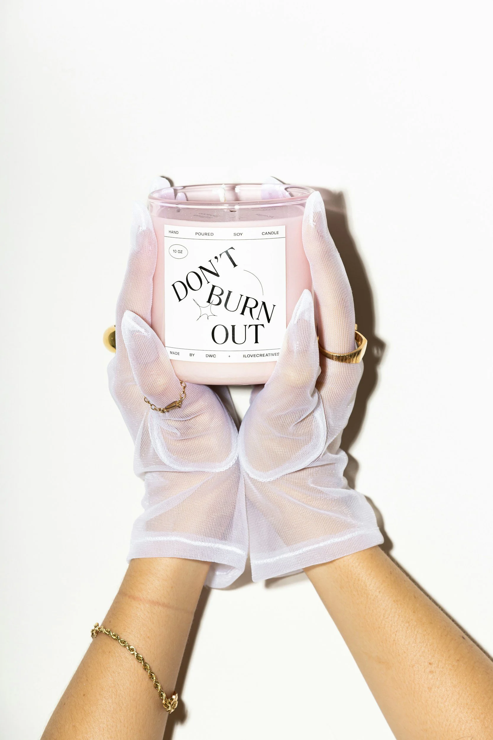 A person wearing white translucent gloves holding a pink candle with a label that says 'Don't Burn Out' on a white background.