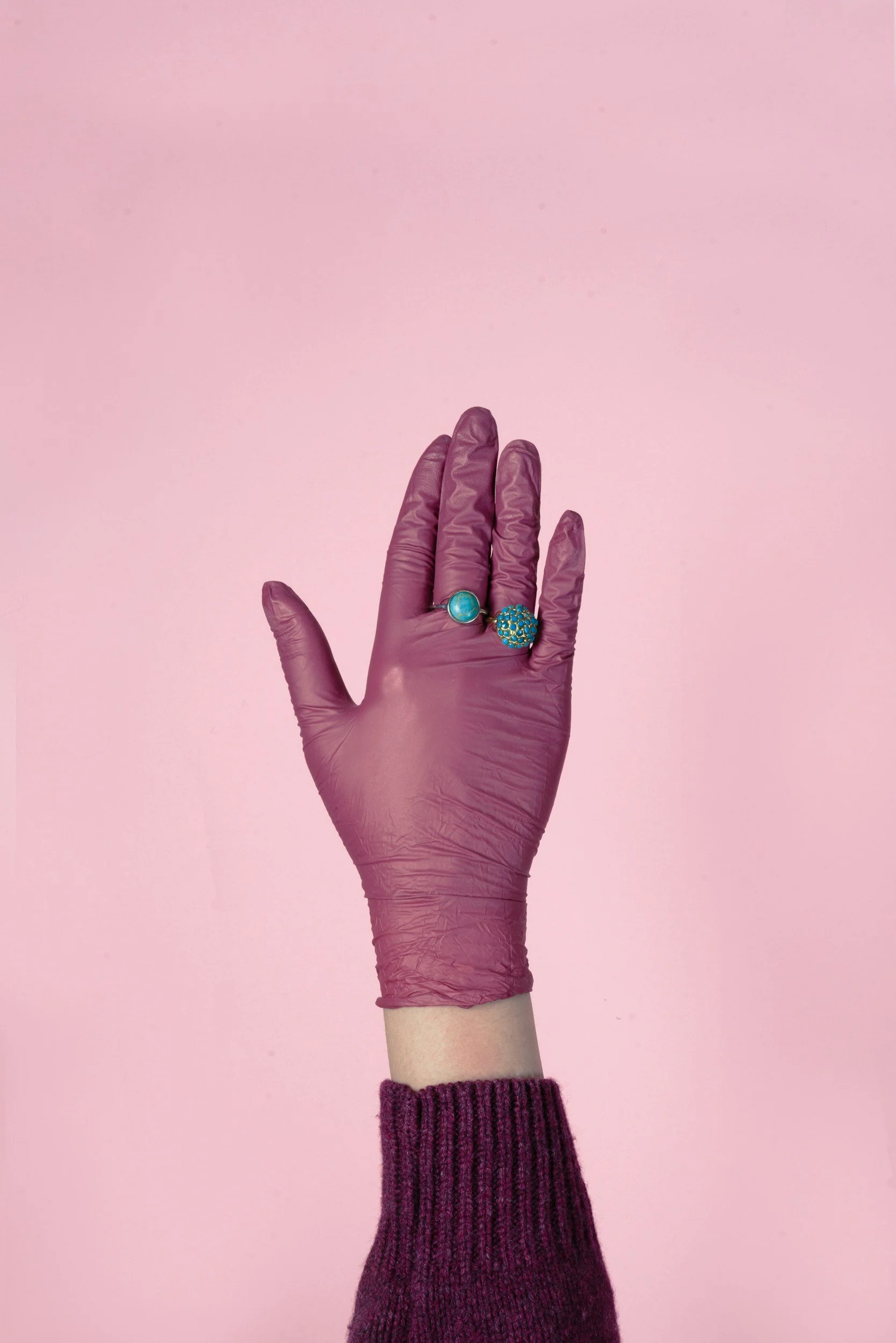 A hand in a purple glove showing two rings against a pink background, with a purple sweater sleeve.