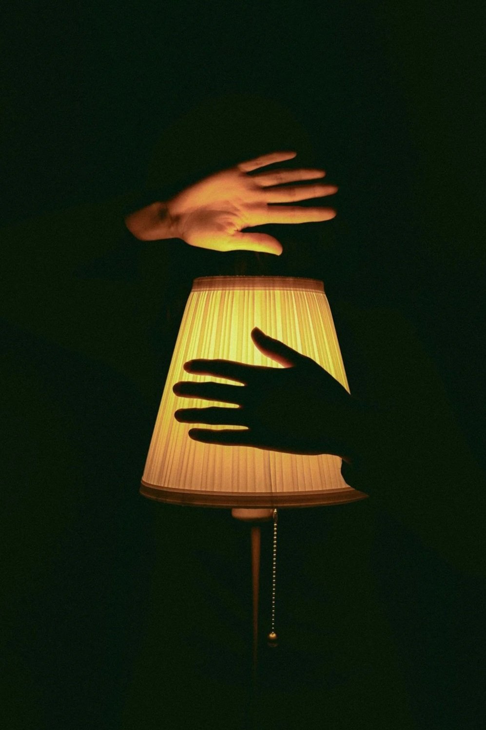 Silhouettes of two hands reaching towards each other with a lamp in the background, creating a dramatic contrast and artistic visual effect.