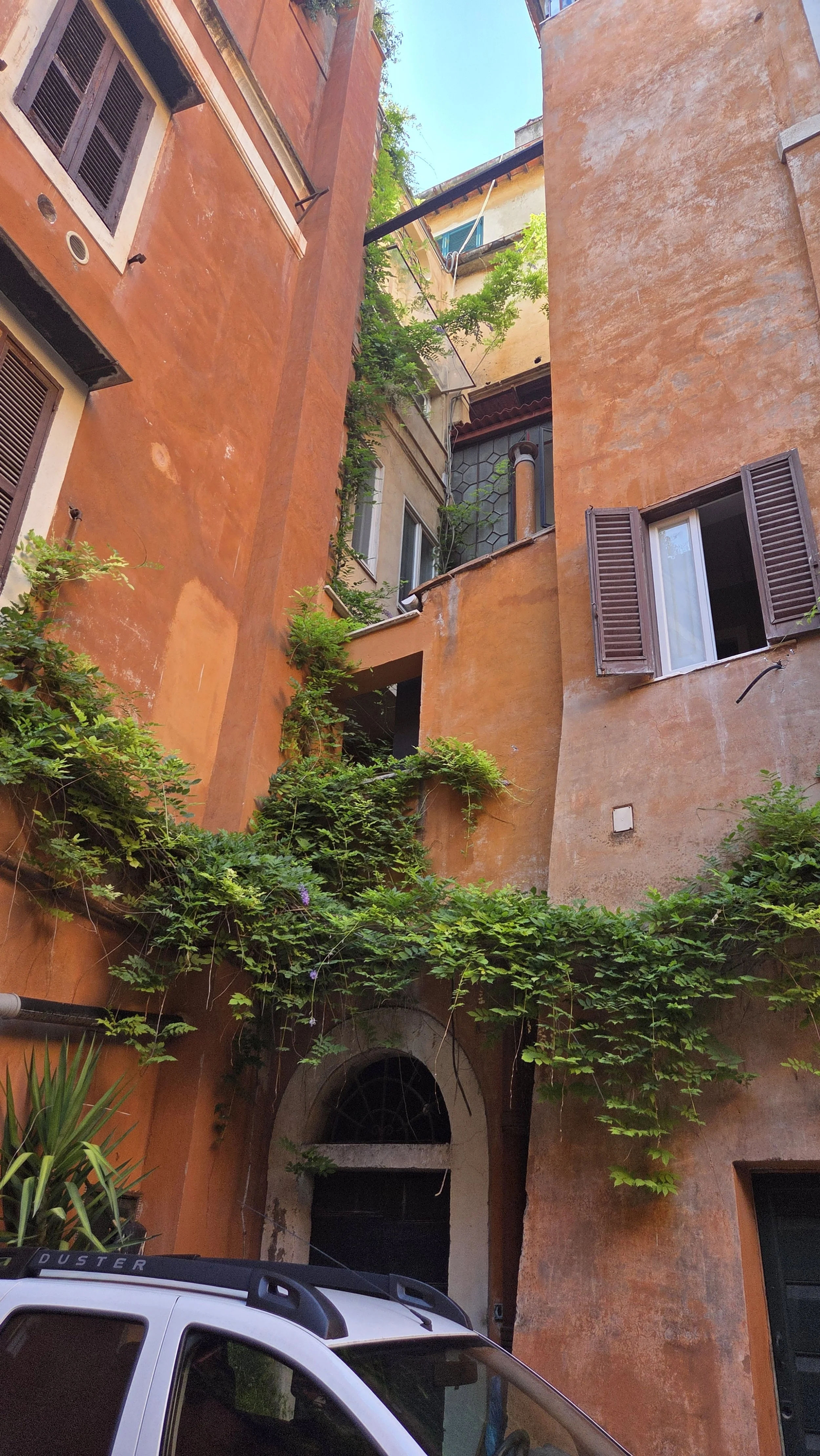 Blick nach oben in einen engen, alten Gebäughof mit orangefarbenen Wänden, grünen Pflanzen und offenen Fenstern.