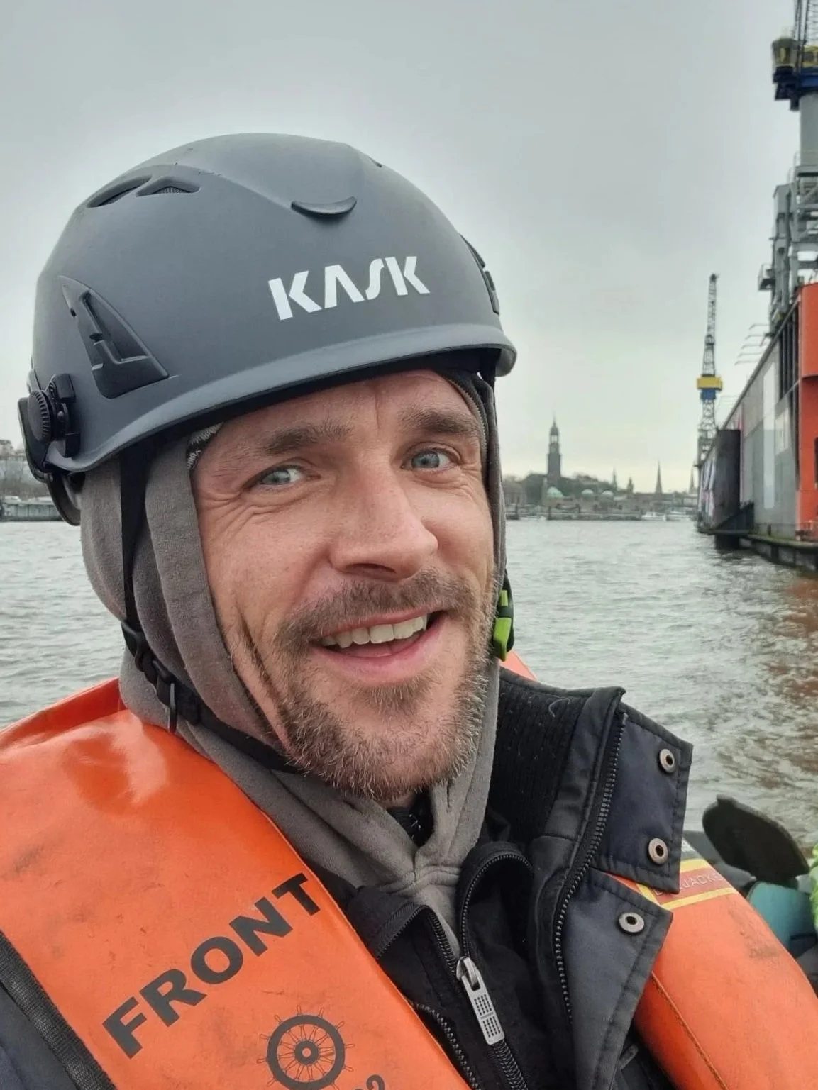 Ein Mann trägt einen Schutzhelm und eine Rettungsweste auf einem Boot auf dem Wasser, mit einer Stadt im Hintergrund unter einem grauen Himmel.