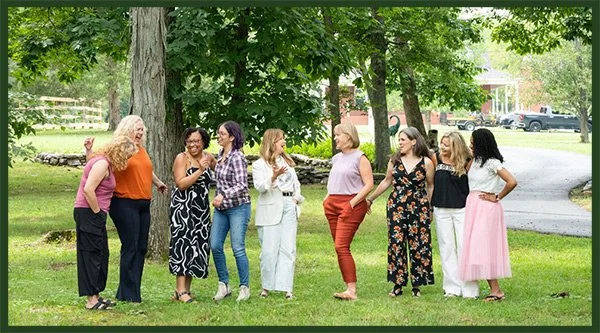 Group of nine women standing on grass under trees in a park near Asheville NC, engaging in conversation and smiling.